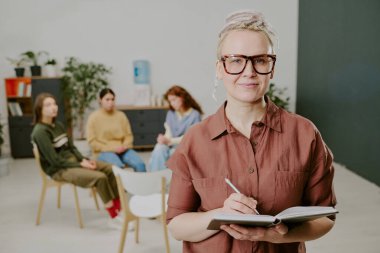 Sınıfta öğrencilerle birlikte dururken elinde defter tutan kadının portresi. Sınıf ortamı etkin öğrenmeyi öneren eğitim araçları ve arkaplan unsurlarını içerir