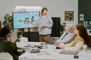 Ekranın yanında duran kadın, arka planda bitki ve güneş paneli olan ofis ortamındaki meslektaşlarına rüzgar türbinlerini açıklıyor. Meslektaşlar özenli ve meşgul görünüyorlar