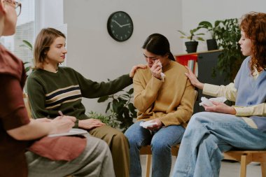 Konforlu bir odada grup terapisi sırasında katılımcılar duygusal destek sağlıyorlar. Bitkiler ve duvarda büyük bir saat var.