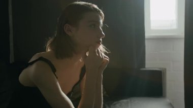 Düşünceli beyaz kadın ellerine bakıyor, sonra yüzünü örtüyor ve saçlarına dokunuyor, koltukta oturuyor, pencereden süzülen soluk bir ışık.