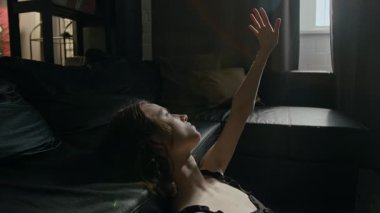 Oturma odasında üzgün bir kız, kafası siyah deri koltukta oturuyor ve kolu pencereye doğru kalkıyor. İçeri ışık giriyor, sonra sağ tarafını çeviriyor.