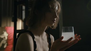 Düşüncelere dalmış siyah gecelik giymiş elinde tabletli bir bardak su tutan genç bir kadın.