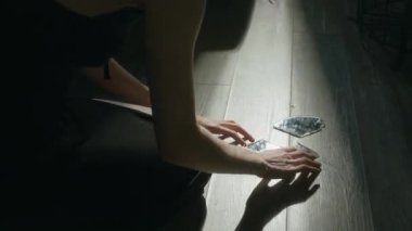 Siyah gecelikli tanınmayan genç bir kadın gergin bir şekilde kırık ayna parçalarına dokunuyor ve onları dairesel bir şekilde yere diziyor.