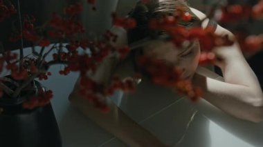 Mutfak masasında beyaz bir vazo dolusu kırmızı böğürtlen ile başını koluna yaslayıp umutsuzluğa kapılmış genç bir kadının karamsar pozunu çek.