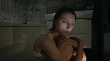 Kafası karışmış, saçları nemli, kameraya bakan beyaz bir kızın portresi. Loş ışıkta, küvetin yanında dizlerinin üzerinde otururken kameraya sarılmış.