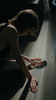 Sabahın erken saatlerinde yerde otururken titreyen ellerle kırık ayna parçalarını bir araya getiren endişeli kızın dikey görüntüsü