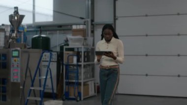 Endüstriyel makinelerin arasında yürüyen ve kahve pişirme tesisinde dijital tablet kullanan ciddi Afrika kökenli Amerikalı kadın yöneticinin yavaş çekimleri.