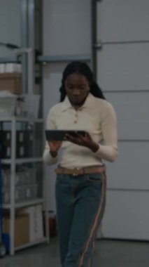 Dijital dizüstü bilgisayar kullanarak ve endüstriyel kahve kızartma tesisine bakarken odaklanmış Afrikalı Amerikalı müdürün dikey portresi.