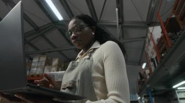 Düşük açılı çekimde, kahve kızartma tesisinde büyük bir depoda dururken, dizüstü bilgisayar kullanan Afrikalı Amerikalı bir işçi görülüyor.