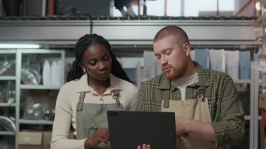 Afro-Amerikalı kadın ve sakallı adam dizüstü bilgisayara odaklanmış, istiflenmiş kutular ve poşetlerin arasında yürüyor kahve kızartma tesisinde