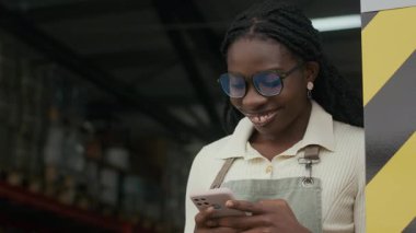 Afrika kökenli bir Amerikalı 'nın önlüklü, gülümseyen, kahve kızartma tesisinde mesai arasında akıllı telefonuyla mesajlaşırken yavaş çekim görüntüsü.