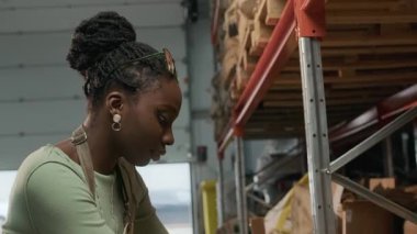 Afrika kökenli Amerikalı kadın işçi önlüğünün eğik çekimleri. Diz çökmüş, çuval bezi tutarken. Dağınık bir kahve deposunda.