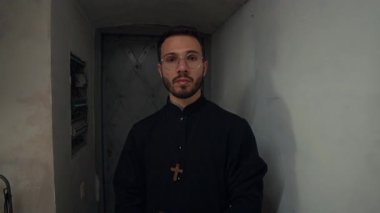 Yüzünde siyah cübbe olan, kilisenin duvarları içinde İncil 'i tutan ve kameraya poz veren kendinden emin genç bir rahibin belinin portresi.