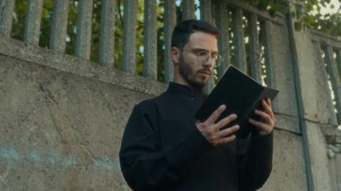 Gözlüklü ve siyah papaz önlüklü ciddi bir rahibin açık havada kutsal İncil okuması.