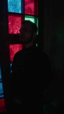 Gözlüklü genç papazın dikey portresi loş ışıkta mozaik camlı pencerenin önünde kameraya poz veriyor.