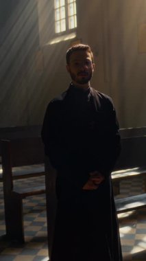 Kilisedeki kameraya siyah cübbe giymiş Orta Doğulu genç Katolik rahibin dikey portresi.