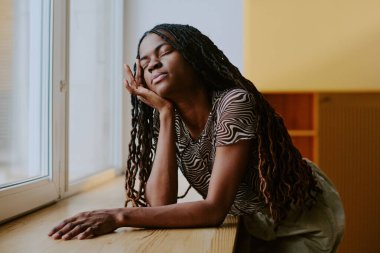 Uzun örgülü kadın pencerenin kenarına yaslanıyor, gözleri kapalı ve huzurlu bir ifadeyle, evde huzurlu bir anın tadını çıkarıyor.