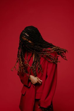 Kırmızı arkaplan önünde duran, uzun dalgalı örgülü, şık kırmızı takım elbiseli, elleri birbirine kenetlenmiş, zarif bir şekilde poz veriyor.