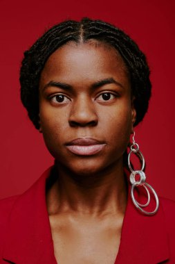 Kırmızı arka planda, çarpıcı dairesel küpeler takan ve kameraya tarafsız bir ifadeyle bakan bir kadının portresi.