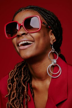 Saçları örgülü, şık güneş gözlüğü takan ve zarif küpeler takan, zengin, kırmızı arka planda kendine güveni ve neşesi olan gülümseyen bir kadının portresi.