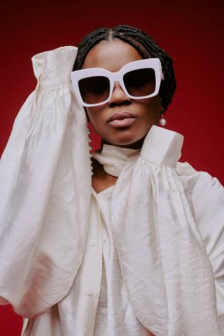 Kabarık kollu beyaz bluz giyen kadın portresi. Katı kırmızı arka planda duruyor ve büyük kare güneş gözlüğü takıyor.