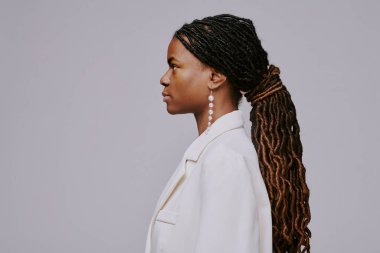 Beyaz ceket giyen, karışık örülmüş saç stili ve uzun küpeler takan bir kadın profili. Yüzünde sakin ve sakin bir ifade