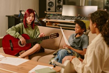 Stüdyoda şarkı ve melodileri birlikte prova eden melez genç grup, akustik gitar tutan ve grup üyeleriyle sohbet eden kızıl saçlı genç kız.