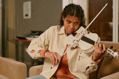 Orta boy, gündelik kıyafetler giymiş, sandalyede oturan ve beyaz keman çalan Afrikalı Amerikalı genç kız şarkı için yeni bir keman rolü yaratıyor.