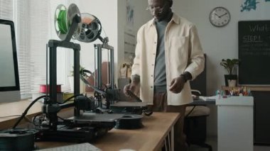 Genç Afrikalı Amerikalı öğretmen telefonunu kontrol ediyor. Modern aydınlık sınıflarda 3D yazıcıların yanında duruyor.