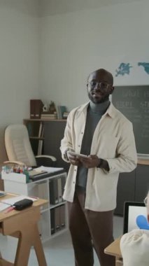 Dikey çekimde genç öğrenciler ellerini kaldırıyor ve Afro-Amerikan erkek öğretmen modern okul sınıfında cevap vermelerine izin veriyor.
