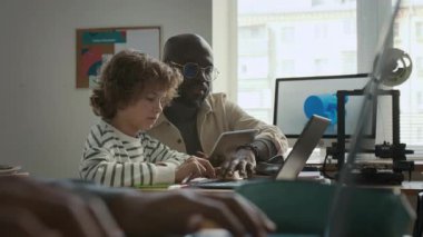 Dost canlısı Afrikalı Amerikalı erkek öğretmen, modern okulda 3D modellik dersi sırasında ilköğretim öğrencisine zorluklar konusunda yardım ediyor.