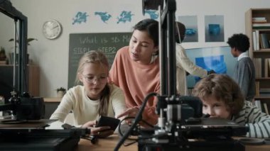 Öğrenciler ve erkek öğretmenler, akıllı telefonlar kullanarak ve sınıfta 3D yazıcı ve karatahta ile 3D model gösteren büyük ekran görüntülerini inceleyerek teknoloji tabanlı proje üzerinde işbirliği yapıyorlar.