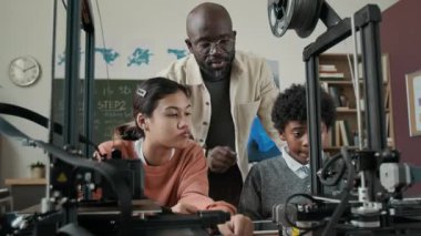 Modern okulda 3Dm modellik dersi sırasında 3D yazıcı kullanarak iki öğrenciye rehberlik eden Afrikalı Amerikalı erkek öğretmen.