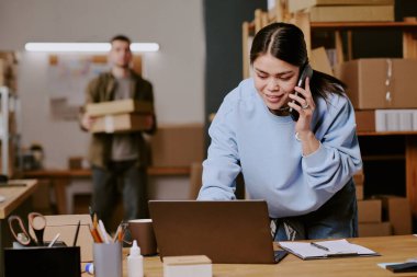 Ofiste dizüstü bilgisayarla yazarken telefonla konuşan bir kadın. Raflar ve arka plandaki kutuları düzenleyen iş arkadaşı
