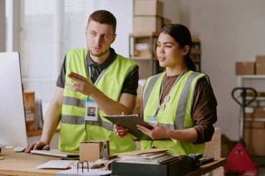 Envanter lojistiğini tartışan iki depo çalışanı dijital tablet ve bilgisayara atıfta bulunurken, arka planda kutularla dolu raflar görülebilir durumda.