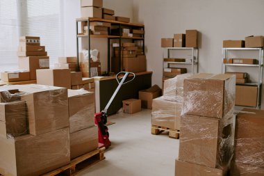 Bir depo ortamında ahşap paletlere ve raflara yığılmış kahverengi karton kutulardan oluşan bir koleksiyon.