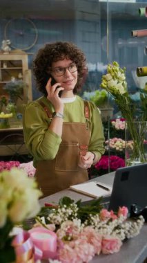 Kıvırcık saçlı, önlüklü, müşteriyle telefonda konuşan ve çiçekçide dizüstü bilgisayarda yazan arkadaş canlısı kadın çiçekçinin dikey görüntüsü.