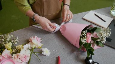 Çiçekçide çiçekçi masasında çiçekçi düzenlemesi yaptıktan sonra beyaz kurdeleyle fiyonk bağlayan...