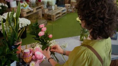 Kıvırcık saçlı kadın çiçekçi dükkanında çalışırken pembe güller ve beyaz çingenelerle çiçek aranjmanı yapıyor.
