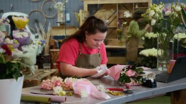 Down sendromlu beyaz esmer çiçekçi. Çiçekçide küçük bir buket gül ve çingene buketinin etrafına beyaz kurdele bağlıyor.