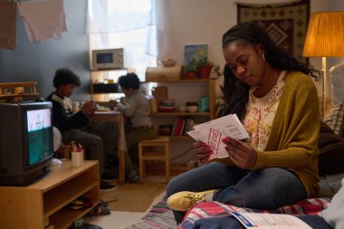 Sıcak aydınlatma ile rahat oturma odasında mektup okuyan bir kadın, arka planda aktivitelerle meşgul olurken, ev ortamını da katıyor.