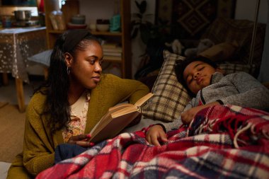 Afrika kökenli Amerikalı anne çocuğuna masal okuyor, çocuk sıcak battaniyenin altında uyurken annesi kitabı tutuyor, rahat bir ortam yaratıyor.