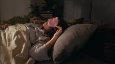 Yuong kadını, bej battaniyeyle örtülmüş ve pembe uyku maskesi takmış, mavi akıllı telefonun yanındaki gri yastıkta sakince uyuyor.
