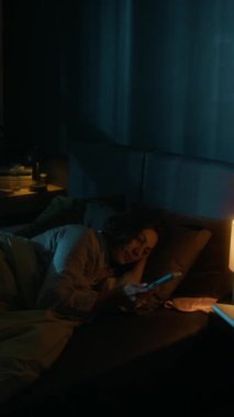 Akıllı telefonunu kilitleyen ve rahat yatağında uyumadan önce ışıkları kapatan genç bir kadının dikey görüntüsü.