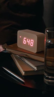 Beyaz dijital alarm saatinin dikey görüntüsü komodinin üzerindeki su bardağının yanındaki yığılmış kitapların üstünde.