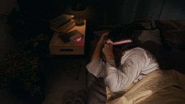 Yüksek açılı, uyku maskeli ve pijamalı bir kadının dijital saat alarmını devre dışı bırakıp tekrar yatağında uyuduğu görüntüler.