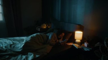Genç bir kadın akıllı telefonunu indiriyor, komodinin ışığını söndürüyor ve modern yatak odasında uyuyakalıyor.
