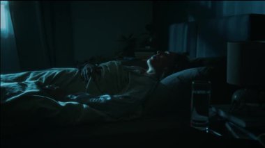Siyah saçlı genç bir kadın geceleri kabustan uyanıyor ve bir bardak su içiyor, yatakta yatıyor.