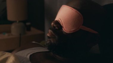 Uykulu Afro-Amerikan bir adamın yakın plan fotoğrafı. Alnına uyku maskesi takmış, uyandıktan sonra yatakta uzanmış.