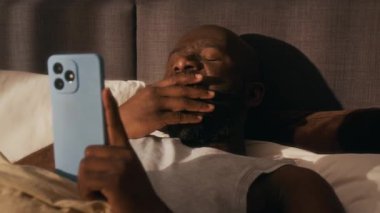 Afro-Amerikalı bir adam sabah güneşli yatak odasında uyandıktan sonra akıllı telefonunu kullanırken esniyor.